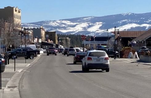 View of a town's main street with buildings on both sides, cars driving through, and mountain covered in snow in the background