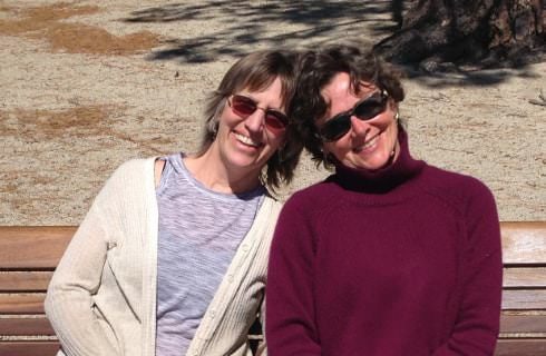 Two women with sunglasses sitting on a wooden bench smiling and looking at the camera with their heads touching