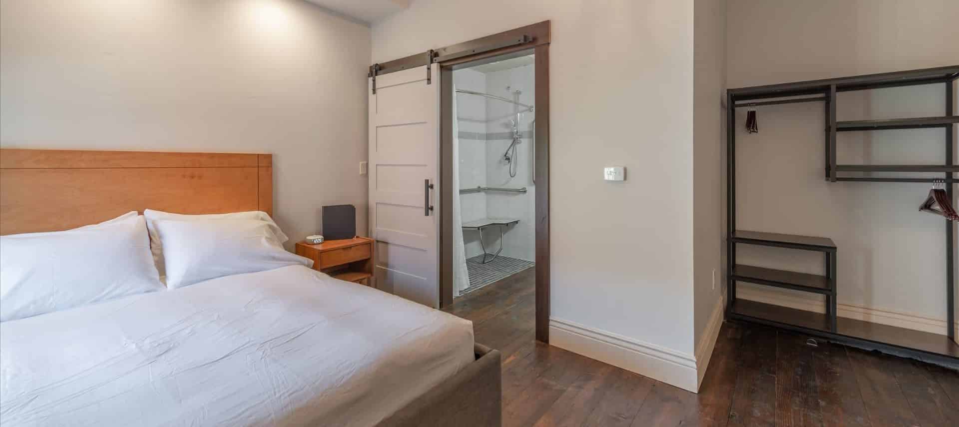 Large bedroom with hardwood floors, white walls, light wooden bed, white bedding, gray metal shelving, and view into bathroom